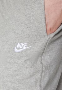 Grå sweatpants i mjuk bomullsblandning, med en broderad vit Nike-logga på sidan, sidofickor och synliga sömnadsdetaljer.
