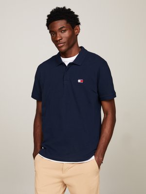 Camisa polo azul marino de algodón con cuello corto, que presenta un pequeño logo en el pecho y se combina con pantalones beige.