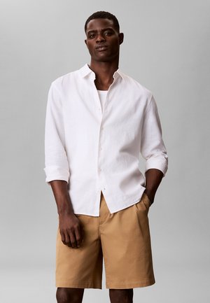 Mężczyzna w białej koszuli z długim rękawem na białym podkoszulku i beżowych szortach do kolan, stoi z rękami w kieszeniach.