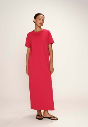 Robe maxi en coton rose vif. Manches courtes, col rond, coupe décontractée. Portée avec des tongs noires. Design simple, texture lisse.