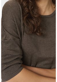 Femme aux cheveux bouclés bruns portant un pull en tricot marron à manches longues, manches remontées, bras croisés à la taille.