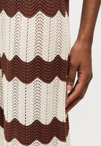 Χέρι που ακουμπά δίπλα σε πλεκτό ύφασμα με εναλλασσόμενες καφέ και λευκές γραμμές σε σχήμα ζιγκ-ζαγκ και ανοιχτό δαντελένιο σχέδιο.