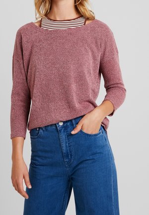 Vrouw draagt een getextureerde mauve trui over een bruin-wit gestreept topje gecombineerd met een high-waisted blauwe spijkerbroek, hand in de zak.