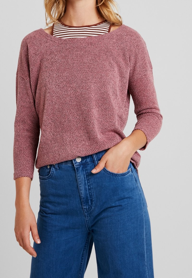 Femme portant un pull mauve texturé sur un haut rayé marron et blanc, associé à un jean bleu taille haute, main dans la poche.