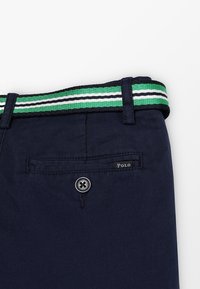 Pantaloni blu navy con una cintura intrecciata a righe verdi e bianche. Presentano una chiusura con bottone e una tasca posteriore con etichetta "Polo".