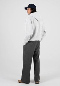 Šedá mikina s kapucí na šňůrku, v kombinaci s tmavě šedými kalhotami s širokými nohavicemi a hnědými botami. Model se otáčí zády proti jednoduchému pozadí.