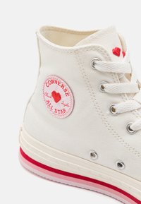 Valge lõuendist kõrge ülemise osaga toss, millel on punane südame logo patch, kontrastne kummist tall ja metallist silmad. Tekstuuriga kangas ja õmblusdetailid.