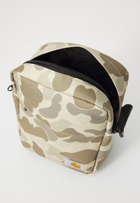 Sac bandoulière en tissu beige à motif camouflage, avec une fermeture éclair noire, un haut ouvert, une poche avant et une petite étiquette logographique en bas à droite.