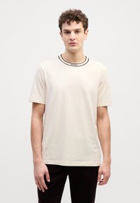T-shirt beige à manches courtes avec un col brodé noir. Texture subtile, coupe décontractée, et présente le nom de la marque "BOSS" au niveau du col.