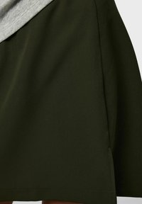 Tumši zaļa svārku ar gludu tekstūru, asimetrisku pelēku augšējo paneli, sānu šķēlumiem un tīru, taisnu apakšmalu.