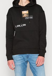 Czarna bluza z kapturem wykonana z bawełny, z grafiką i napisem na przodzie. Luźny krój, kieszeń typu kangurek oraz ściągacze na rękawach. W zestawie z jasnoniebieskimi dżinsami.