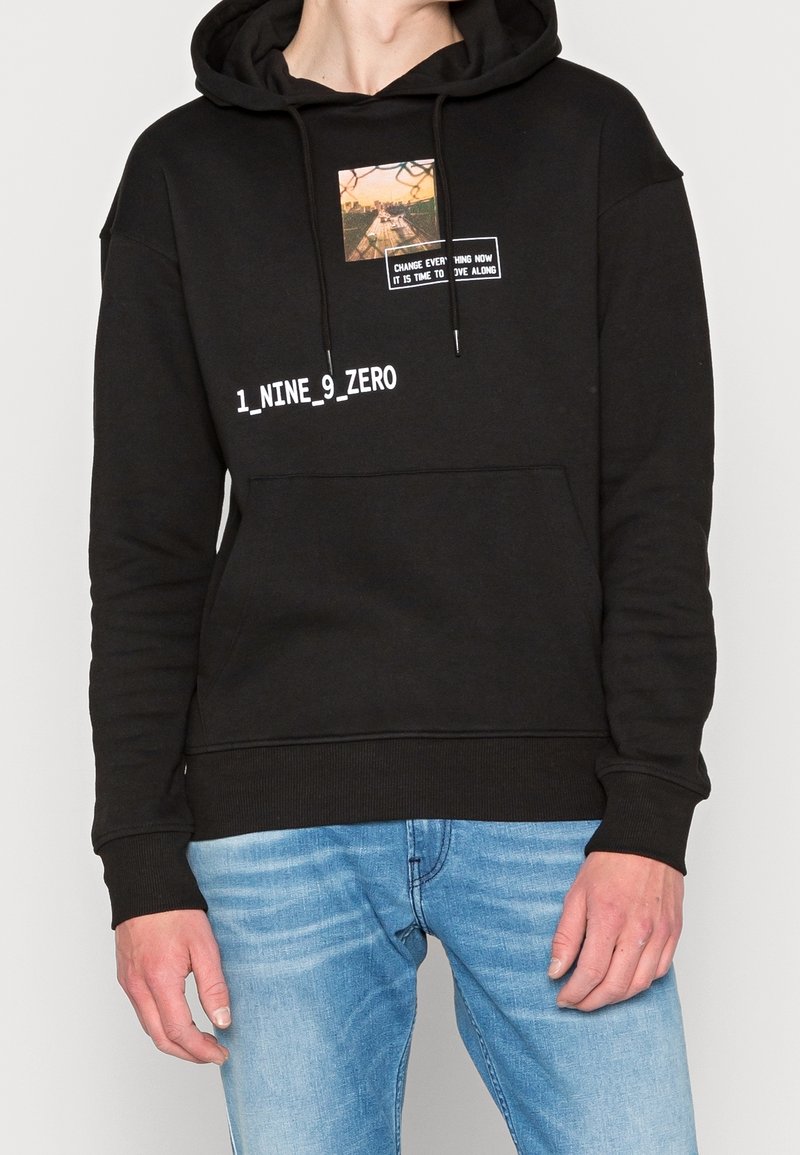 Sudadera con capucha negra de algodón, con un gráfico y texto en la parte frontal. Corte holgado, bolsillo tipo canguro y puños acanalados. Combinada con jeans azul claro.