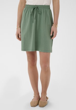 Kvinde iført et knælængde grønt skørt med elastisk talje og snor, kombineret med beige vævede flade sko, står mod en hvid baggrund.