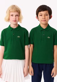 Gröna polo skjortor med korta ärmar, med en placket med tre knappar och en liten logotyp. Flickan bär en vit plisserad kjol, pojken mörka byxor.