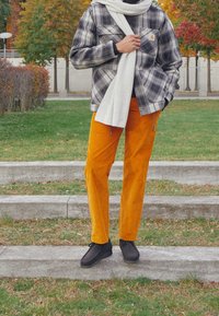 Karierte Flanelljacke in Grau und Schwarz, kombiniert mit orangefarbigen Cordhosen. Eingewickelt in einen hellgrauen Schal und trägt schwarze Schuhe.
