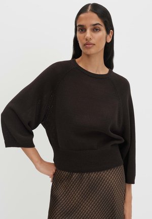 Femme aux longs cheveux foncés porte un pull noir ample à manches larges et une jupe marron à motifs, posant avec la main sur la hanche sur un fond uni.