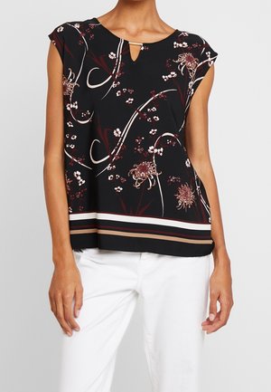 Top noir sans manches à imprimé floral et encolure en forme de goutte, associé à un pantalon blanc porté par une personne debout sur un fond uni.