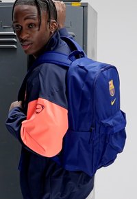 Marinblå ryggsäck med mjuk textur, försedd med en Barcelona-logotyp och en ficka med dragkedja. Bärs över ena axeln, kombinerad med en sportig jacka.