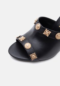 Μαύρο mule με ανοιχτή μύτη και δύο λουριά που διαθέτουν χρυσά διακοσμητικά καρφάκια σε σχήμα κύκλου και λουλουδιού.