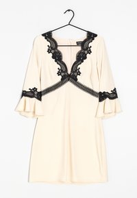 Krämfärgad klänning med svarta spetsdetaljer, V-ringning med blommönster, klockärmar och åtsittande silhuett, hängande på en svart krok.