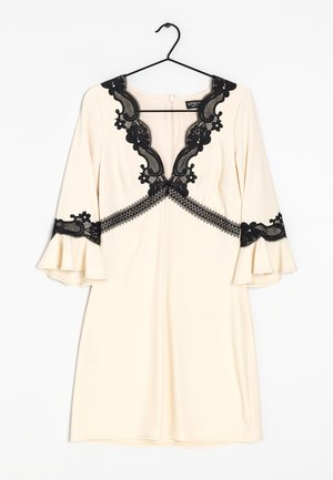 Cremefarbenes Kleid mit schwarzen Spitzenakzenten, V-Ausschnitt mit floralem Muster, Glockenärmel und taillierte Silhouette, hängt an einem schwarzen Haken.
