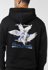 Mann trägt schwarzen Kapuzenpullover mit weißen Tauben und blau-weißem "Closure London"-Text auf der Rückseite vor einfachem Hintergrund.