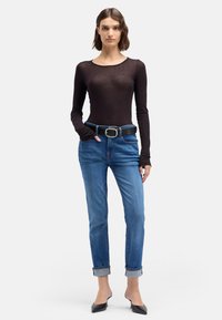 Haut noir à manches longues en tissu léger, associé à un jean bleu retroussé et une ceinture noire avec une grande boucle argentée. Chaussures noires à bouts pointus.