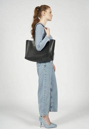 Borsa a tracolla in pelle nera con cuciture verticali, forma rettangolare e manici lunghi, tenuta da una persona con una maglietta a righe blu e jeans.