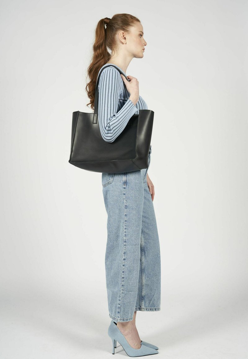 Czarna skórzana torba typu tote z pionowymi szwami, prostokątnym kształtem i długimi uchwytami, trzymana przez osobę w niebiesko-paskowym topie i dżinsach.