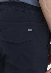 Navy blue fabric shorts with an elastic waistband, featuring a back pocket and a small striped accent tag. Smooth texture, casual design.