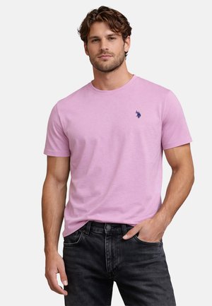 Mann trägt ein rosa kurzärmeliges T-Shirt mit kleinem marineblauem Logo auf der Brust und schwarze Jeans, Hand in der Tasche.