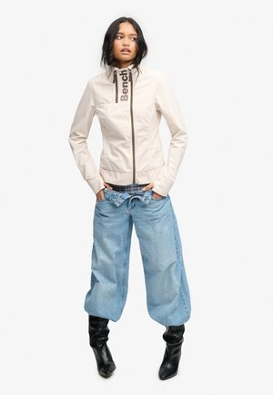 Ung kvinde med langt mørkt hår iført en lysebeige lynlåsjakke, løstsiddende lyseblå jeans og sorte støvler, stående med hænderne i lommerne.