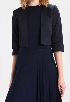 Frau trägt eine marineblaue, kurz geschnittene Jacke mit dreiviertellangen Ärmeln über einem plissierten, marineblauen Kleid vor einfachem Hintergrund.