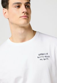 Valkoinen puuvillapaita lyhyillä hihoilla, jossa on mustalla brodeeratulla tekstillä varustettu vasen rinta-alue: "WRANGLER BUTTS DRIVE ME NUTS".