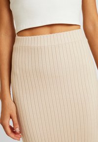 Beżowa midi spódnica z prążkowanego dzianiny z wysokim elastycznym pasem, z pionowymi paskami i miękką teksturą. Zbliżenie na spódnicę i rękę.