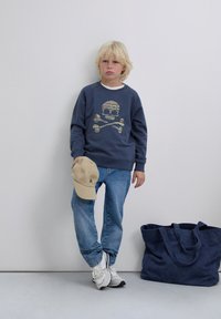 Un niño rubio está de pie contra la pared, vistiendo un sudadera azul con un estampado de calaveras, jeans, zapatillas blancas, sosteniendo una gorra beige, con una bolsa azul marino en el suelo.