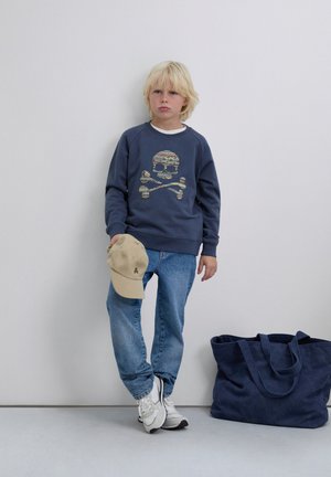 Blond child stands against wall wearing blue skull-patterned sweatshirt, jeans, white sneakers, holding beige cap, with navy bag on floor.