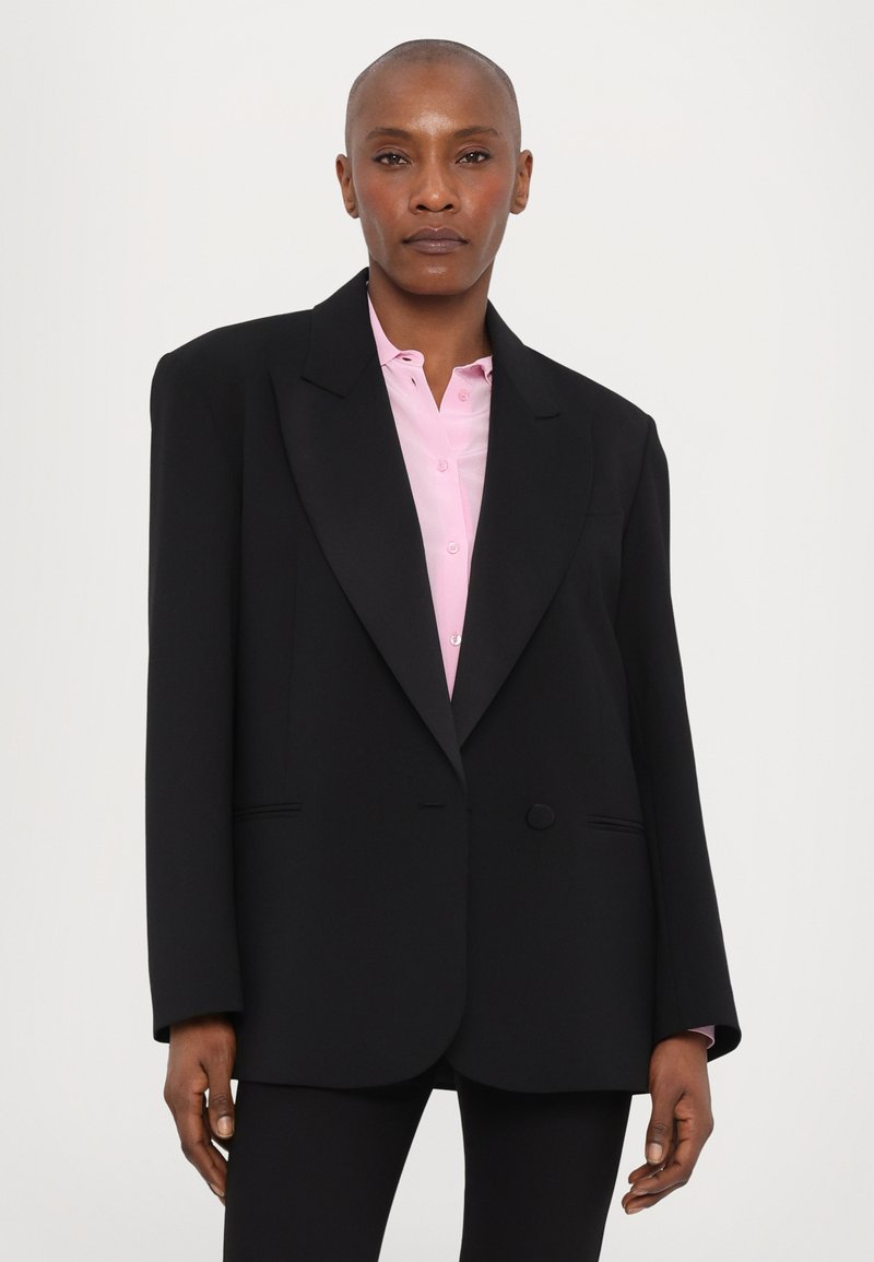 Czarna marynarka o dopasowanym kroju, szerokich klapach i z jednym zapięciem na guzik. Noszona na jasnoróżową koszulę, gładki materiał, z dwiema przednimi kieszeniami.