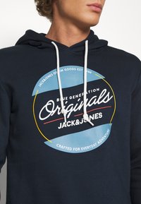 Marinblå hoodie med justerbara snoddar, prydd med en tryckt cirkellogo i blått, gult och vitt. Mjuk tygkvalitet, avslappnad design.