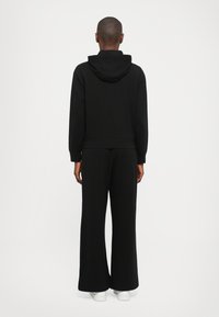 Čierna mikina s kapucňou a široké nohavice, vyrobené z jemného materiálu. Obsahuje kapucňu a rebrované náplety. Biele tenisky dopĺňajú outfit.
