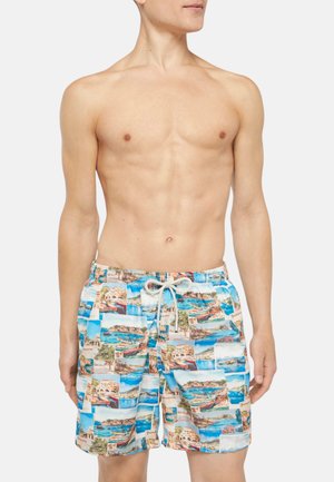 Man zonder shirt die blauwe zwembroek draagt met foto-collage van een schilderachtige kust en boot en trekkoort taille, staand tegen een witte achtergrond.