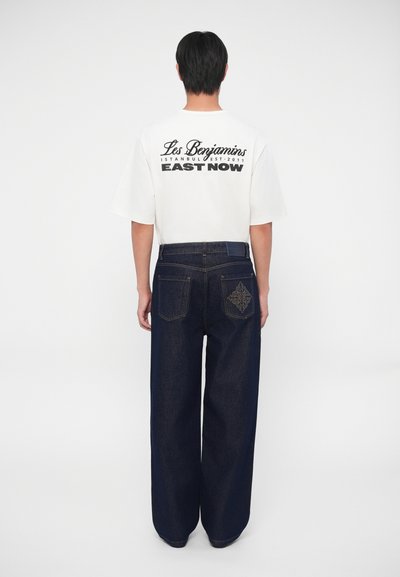 Camiseta oversized blanca con letras negras en la parte posterior. Jeans de mezclilla de pierna ancha azul oscuro con costuras decorativas en el bolsillo trasero.