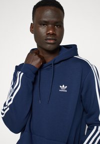 Marinblå Adidas hoodie med en framficka i kängurustil, vita accentremsor på ärmarna och en liten vit logotyp på bröstet. Mjuk textur.