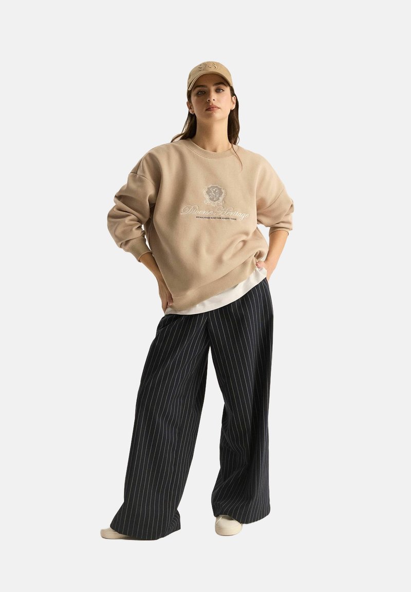 Beżowa, oversize'owa bluza z haftowanym logo, zestawiona z czarnymi spodniami w szerokie paski, oraz beżową czapką. Widoczna biała warstwowa koszula.