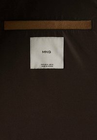 Tessuto marrone con una texture liscia, caratterizzato da un'etichetta bianca rettangolare con "MNG" e dettagli sulle cure, e una striscia marrone a contrasto sopra.