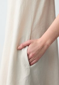 Mano che riposa all'interno della tasca laterale di un indumento in lino beige chiaro e morbido, su uno sfondo semplice e chiaro.