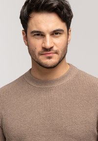 Brązowy sweter dzianinowy z okrągłym dekoltem. Tkanina ma teksturowany wzór i jest noszona przez mężczyznę z krótkimi ciemnymi włosami i zarostem na twarzy.