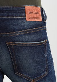 Baksidan av mörkblå denimjeans med orange sömmar och en brun läderetikett märkt "ONLY & SONS" ovanför den högra bakfickan.