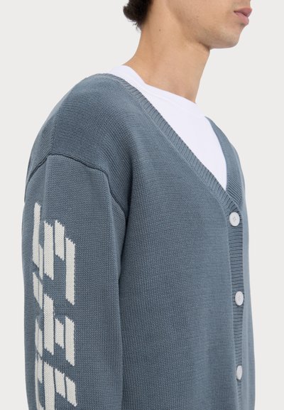 Šedý pletený kardigán s výstrihom do V, so zapínaním na biele gombíky a geometrickým vzorom na ľavom rukáve. Hladká textúra.