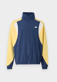 Veste zippée bleu marine et jaune avec col haut, présentant un design color-block et des poignets élastiques ; le tissu semble lisse et sportif.